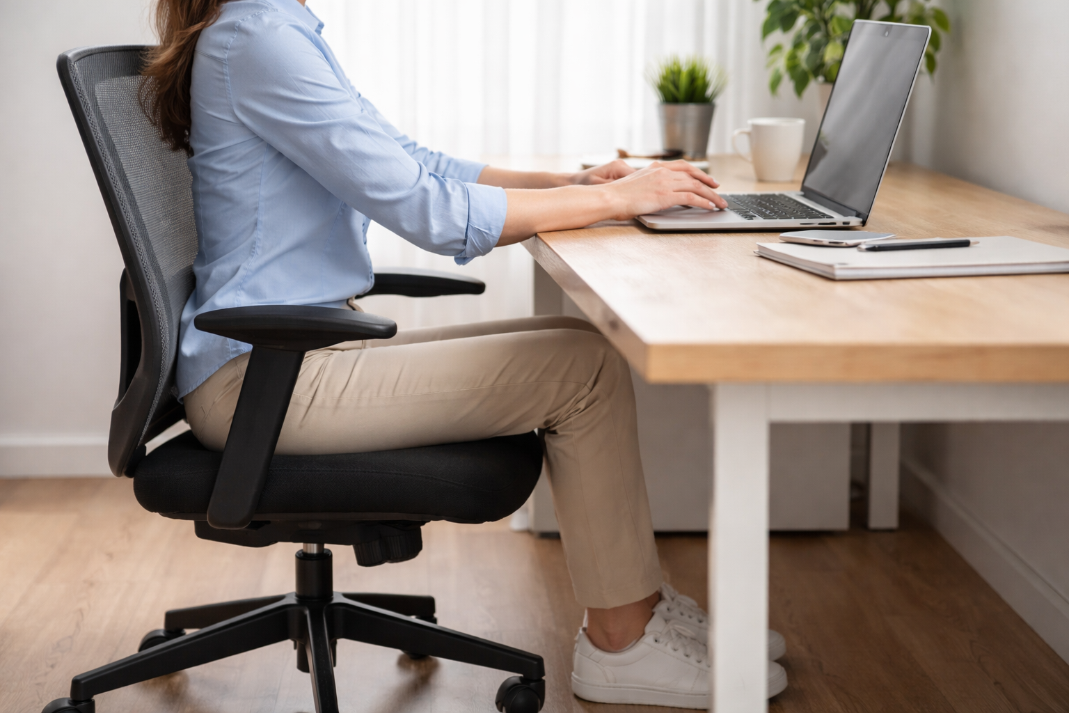 Boczny kadr osoby siedzącej ergonomicznie przy biurku: plecy oparte o oparcie, stopy na podłodze, przedramiona podparte na blacie, praca przy laptopie.